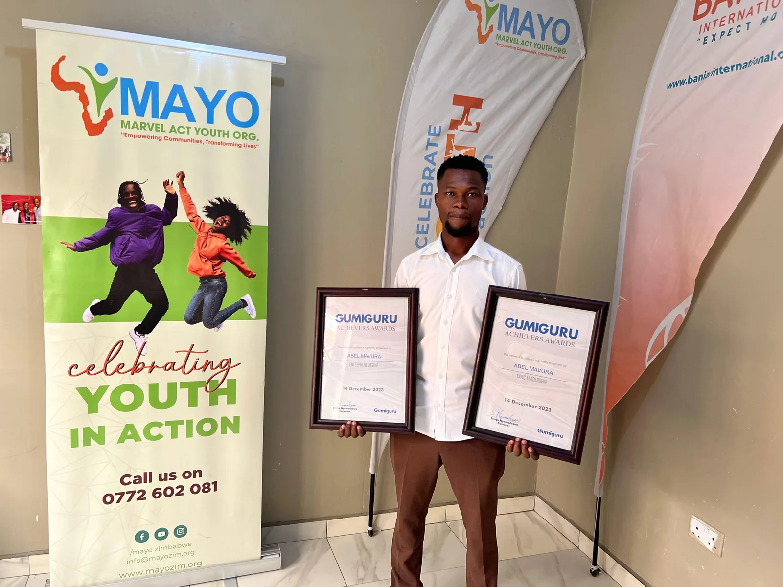 Abel Mavura holding two Gumiguru award certificates in front of a MAYO banner